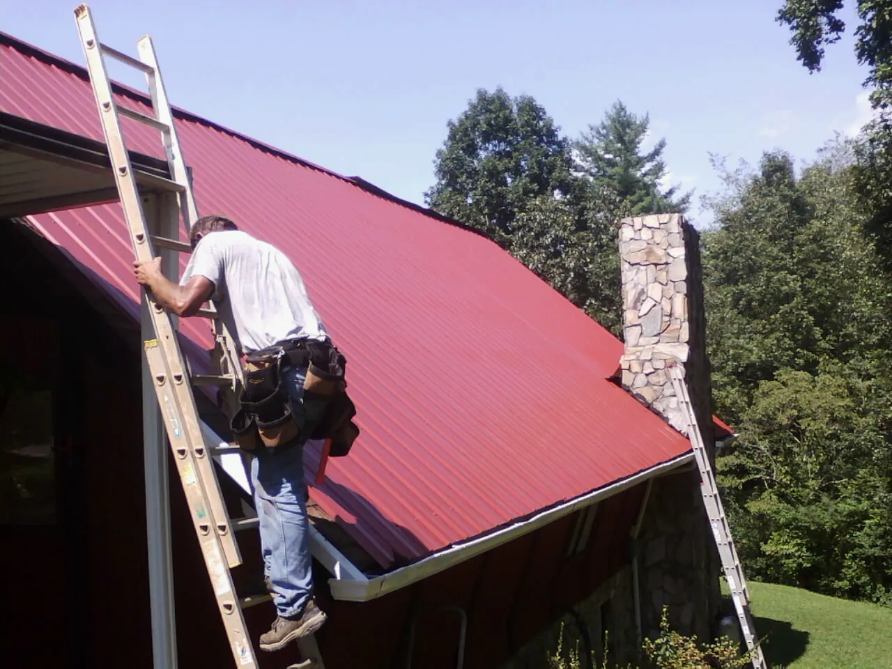 Roofer on a ladder beside a red metal roof and stone chimney — Soffit & Fascia Repair by Roofing Brothers in Wallpack Center